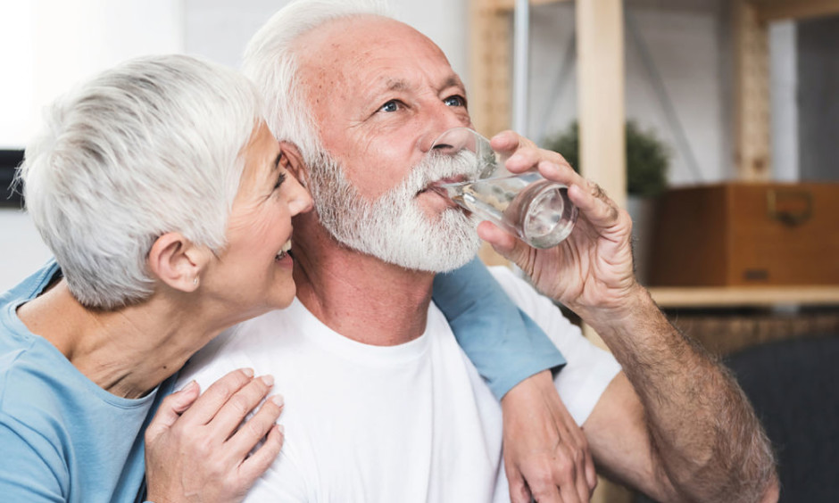Hombre adulto mayor bebiendo un vaso de agua mientras su esposa lo abraza
