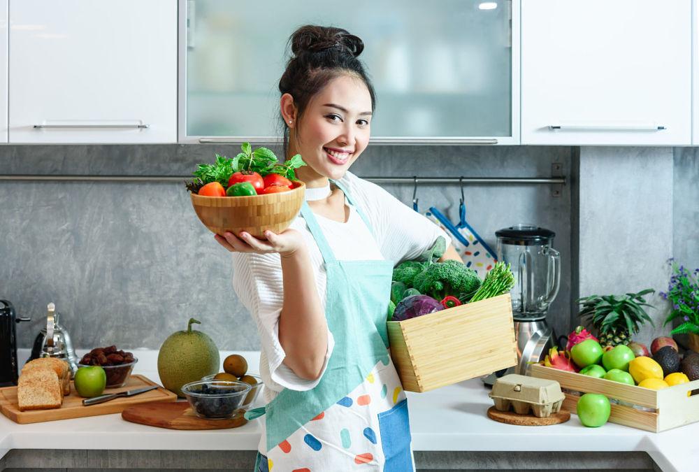 mujer en la cocina sosteniendo varios vegetales y frutas en sus brazos